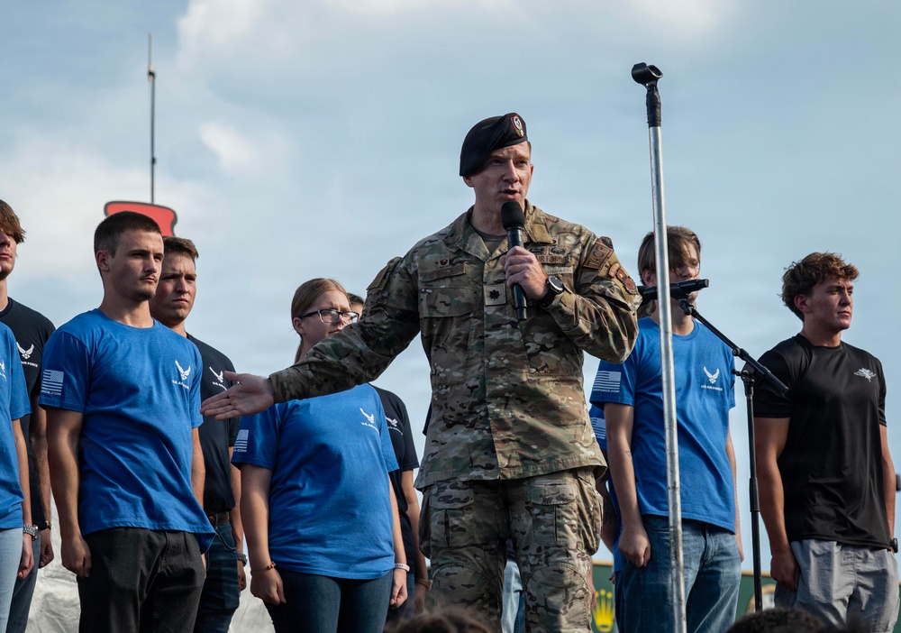 A U.S. Army officer in multicam/OCP uniform and black beret is speaking into a microphone at what appears to be a public event or ceremony. He is standing on a stage surrounded by young people wearing blue and black t-shirts featuring the U.S. Air Force logo. The setting appears to be an outdoor venue with a partly cloudy sky in the background.