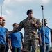 A U.S. Army officer in multicam/OCP uniform and black beret is speaking into a microphone at what appears to be a public event or ceremony. He is standing on a stage surrounded by young people wearing blue and black t-shirts featuring the U.S. Air Force logo. The setting appears to be an outdoor venue with a partly cloudy sky in the background.
