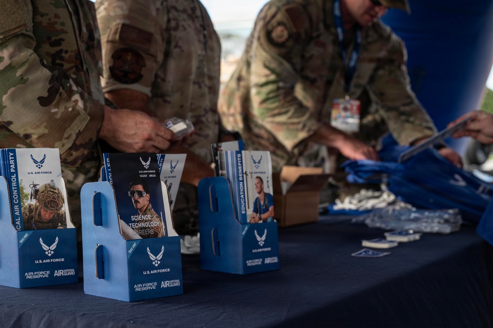 Two U.S. Air Force personnel in OCP (Operational Camouflage Pattern) uniforms are setting up or manning a recruitment/information booth. The table displays multiple blue brochure holders containing U.S. Air Force recruitment pamphlets covering topics such as 'Science &amp; Technology Careers,' 'Education Benefits,' and 'Tactical Air Control Party.' The booth represents a joint recruitment effort for the U.S. Air Force, Air Force Reserve, and Air National Guard.
