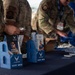 Two U.S. Air Force personnel in OCP (Operational Camouflage Pattern) uniforms are setting up or manning a recruitment/information booth. The table displays multiple blue brochure holders containing U.S. Air Force recruitment pamphlets covering topics such as 'Science &amp; Technology Careers,' 'Education Benefits,' and 'Tactical Air Control Party.' The booth represents a joint recruitment effort for the U.S. Air Force, Air Force Reserve, and Air National Guard.