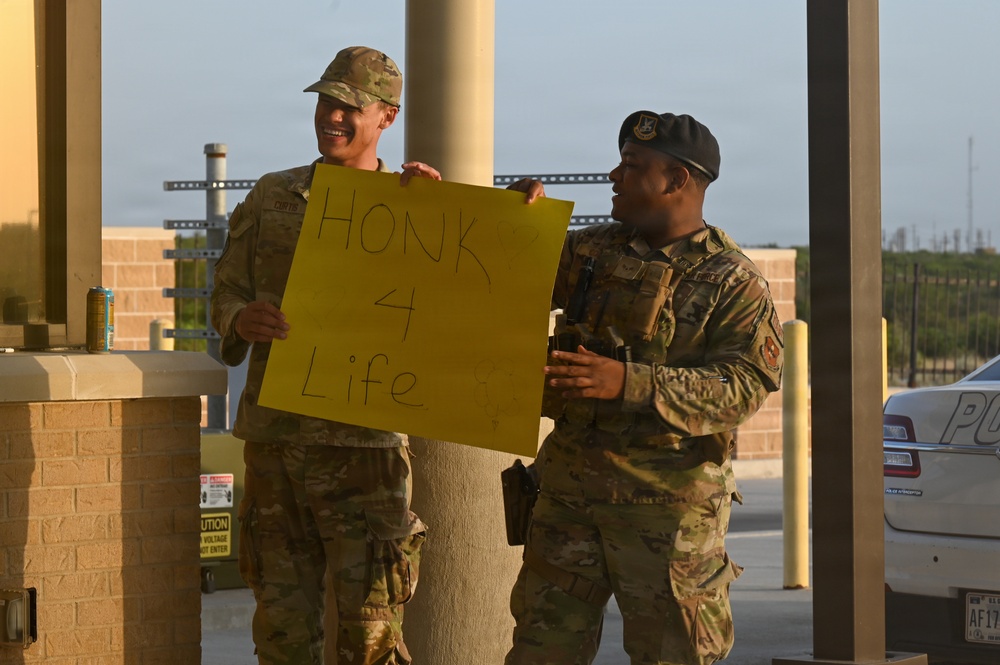 Two military personnel in camouflage uniforms are standing at what appears to be a base entry checkpoint or gate area. One soldier is holding a yellow sign reading 'HONK 4 Life' while both appear to be smiling and in good spirits. The setting suggests a suicide prevention or morale awareness campaign at a military installation.