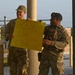 Two military personnel in camouflage uniforms are standing at what appears to be a base entry checkpoint or gate area. One soldier is holding a yellow sign reading 'HONK 4 Life' while both appear to be smiling and in good spirits. The setting suggests a suicide prevention or morale awareness campaign at a military installation.