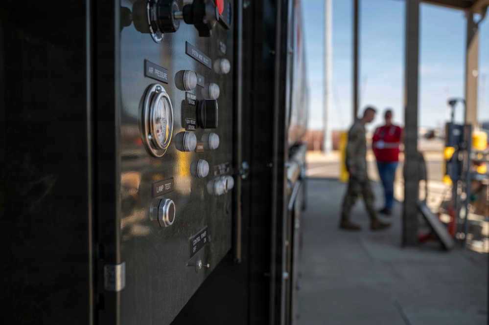 The image shows a close-up of an industrial or military equipment control panel with various gauges, knobs, and buttons in the foreground. In the blurred background, two individuals — one in military camouflage uniform and one in civilian clothing with a red shirt — are standing near what appears to be a fueling or utility station. The setting appears to be an outdoor military or industrial facility under clear skies.