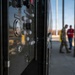 The image shows a close-up of an industrial or military equipment control panel with various gauges, knobs, and buttons in the foreground. In the blurred background, two individuals — one in military camouflage uniform and one in civilian clothing with a red shirt — are standing near what appears to be a fueling or utility station. The setting appears to be an outdoor military or industrial facility under clear skies.