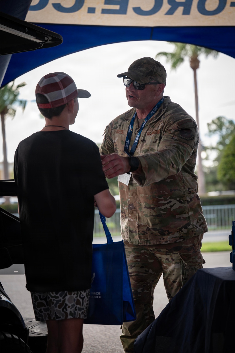 A U.S. Air Force service member in OCP (Operational Camouflage Pattern) uniform is engaged in conversation with a young person at what appears to be a military recruiting or outreach booth. The interaction takes place under a blue canopy tent bearing partial text reading 'FORCE.C' (likely AIRFORCE.COM). The setting appears to be an outdoor public event, possibly a fair or community gathering in a warm climate.