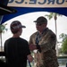 A U.S. Air Force service member in OCP (Operational Camouflage Pattern) uniform is engaged in conversation with a young person at what appears to be a military recruiting or outreach booth. The interaction takes place under a blue canopy tent bearing partial text reading 'FORCE.C' (likely AIRFORCE.COM). The setting appears to be an outdoor public event, possibly a fair or community gathering in a warm climate.