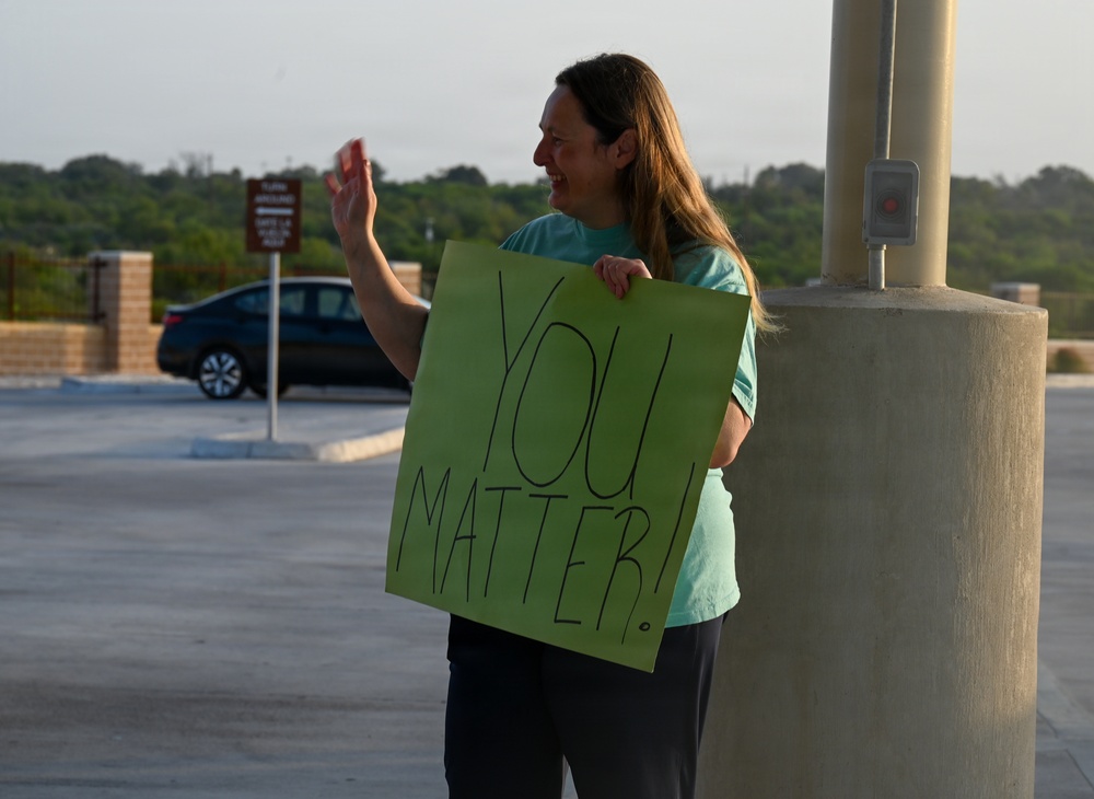A woman stands in what appears to be a parking lot holding a green handmade sign reading 'YOU MATTER!' while waving with her right hand. She is smiling and appears to be greeting or acknowledging passing vehicles or people. The setting appears to be outdoors during early morning or evening based on the warm, low-angle lighting.