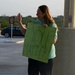 A woman stands in what appears to be a parking lot holding a green handmade sign reading 'YOU MATTER!' while waving with her right hand. She is smiling and appears to be greeting or acknowledging passing vehicles or people. The setting appears to be outdoors during early morning or evening based on the warm, low-angle lighting.