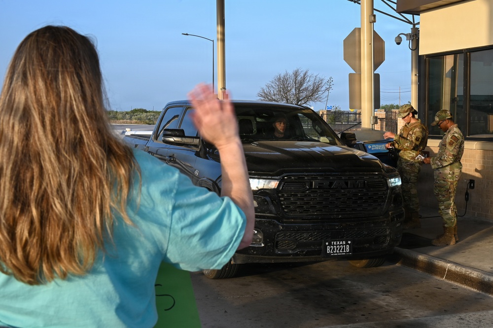 A woman in a teal shirt is waving at the driver of a black Ram pickup truck at what appears to be a military base checkpoint in Texas. Two soldiers in OCP (Operational Camouflage Pattern) uniforms are checking the vehicle's credentials at the guard post. The scene suggests a farewell or greeting moment at a military installation gate.