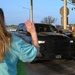 A woman in a teal shirt is waving at the driver of a black Ram pickup truck at what appears to be a military base checkpoint in Texas. Two soldiers in OCP (Operational Camouflage Pattern) uniforms are checking the vehicle's credentials at the guard post. The scene suggests a farewell or greeting moment at a military installation gate.