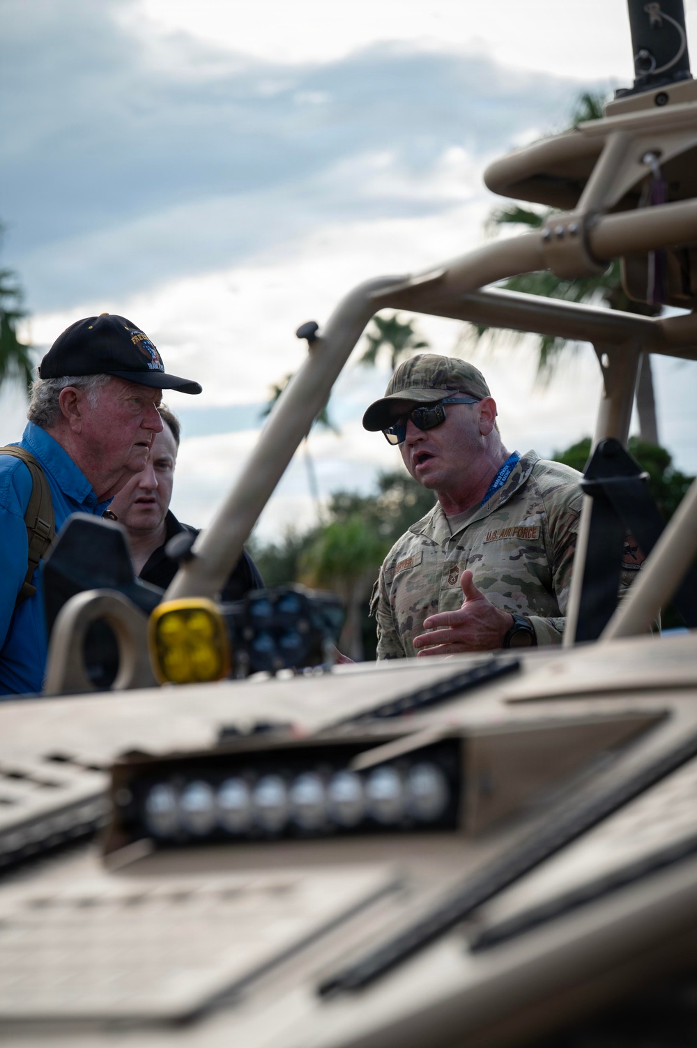 A U.S. Air Force servicemember in OCP (Operational Camouflage Pattern) uniform is briefing or explaining something to two civilians near a military tactical vehicle. The setting appears to be an outdoor demonstration or public event, suggested by the blue lanyards/badges visible on the participants. Palm trees and a cloudy sky are visible in the background.