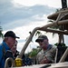 A U.S. Air Force servicemember in OCP (Operational Camouflage Pattern) uniform is briefing or explaining something to two civilians near a military tactical vehicle. The setting appears to be an outdoor demonstration or public event, suggested by the blue lanyards/badges visible on the participants. Palm trees and a cloudy sky are visible in the background.