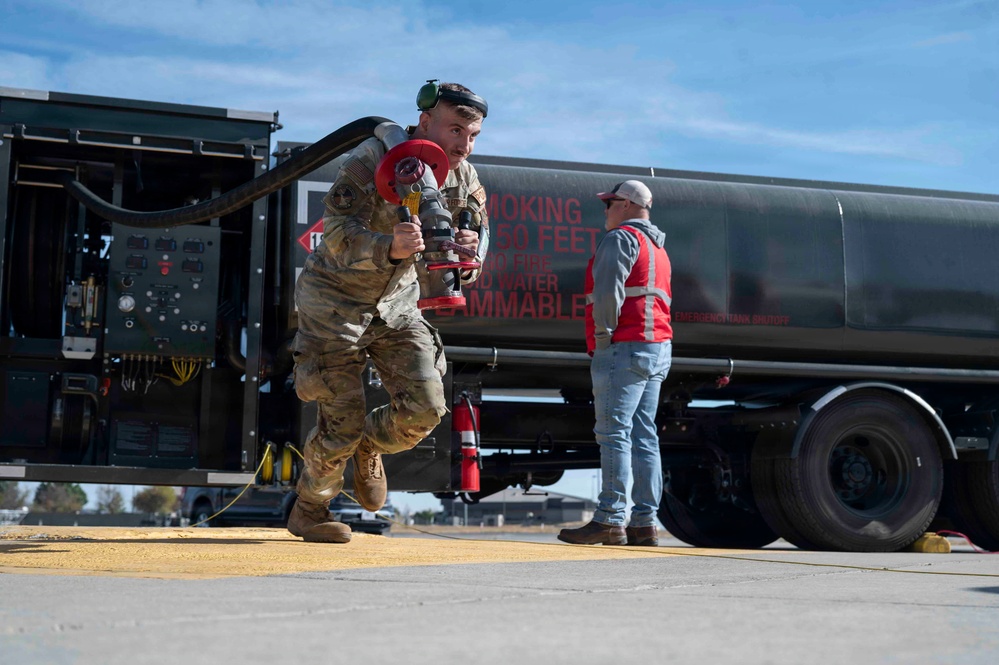 A military service member in camouflage uniform carries a fuel nozzle/coupling assembly near a large dark-colored fuel tanker truck on what appears to be an airfield tarmac. A civilian observer in a red safety vest and white cap stands near the rear of the tanker. The scene depicts a fueling operation or training exercise at a military airfield.