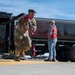 A military service member in camouflage uniform carries a fuel nozzle/coupling assembly near a large dark-colored fuel tanker truck on what appears to be an airfield tarmac. A civilian observer in a red safety vest and white cap stands near the rear of the tanker. The scene depicts a fueling operation or training exercise at a military airfield.