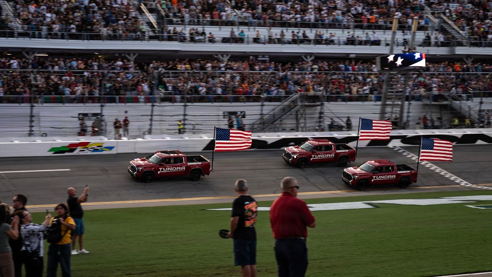 Three red Toyota Tundra pickup trucks drive in formation along a NASCAR racetrack, each carrying an American flag. The event appears to be a pre-race ceremony or parade lap at a major speedway with packed grandstands. Spectators line the infield area watching the procession.