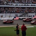 Three red Toyota Tundra pickup trucks drive in formation along a NASCAR racetrack, each carrying an American flag. The event appears to be a pre-race ceremony or parade lap at a major speedway with packed grandstands. Spectators line the infield area watching the procession.