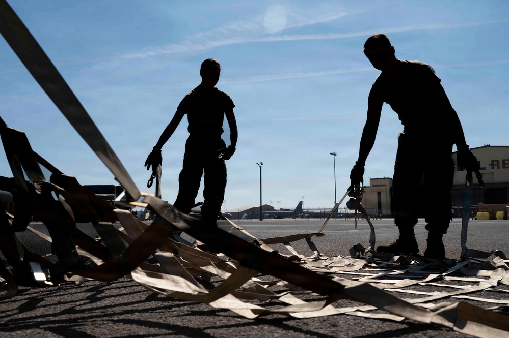 Two silhouetted individuals stand on a military airfield tarmac, handling what appears to be cargo straps or tie-down equipment spread across the ground. The strong backlighting creates dramatic silhouettes against a clear blue sky, with military aircraft and hangar structures visible in the background. The setting appears to be a military air base during daytime operations.