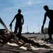 Two silhouetted individuals stand on a military airfield tarmac, handling what appears to be cargo straps or tie-down equipment spread across the ground. The strong backlighting creates dramatic silhouettes against a clear blue sky, with military aircraft and hangar structures visible in the background. The setting appears to be a military air base during daytime operations.