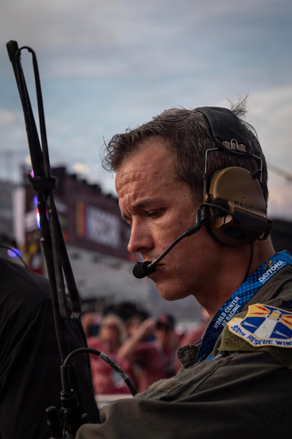 A focused military personnel member wearing a tactical headset with boom microphone is photographed in profile at Daytona International Speedway. He is wearing an olive drab flight suit with a '920th Rescue Wing' patch and a 'Daytona World Center of Racing' credential lanyard. The background shows the grandstands and track environment, suggesting a ceremonial or operational presence at a NASCAR event.