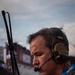 A focused military personnel member wearing a tactical headset with boom microphone is photographed in profile at Daytona International Speedway. He is wearing an olive drab flight suit with a '920th Rescue Wing' patch and a 'Daytona World Center of Racing' credential lanyard. The background shows the grandstands and track environment, suggesting a ceremonial or operational presence at a NASCAR event.