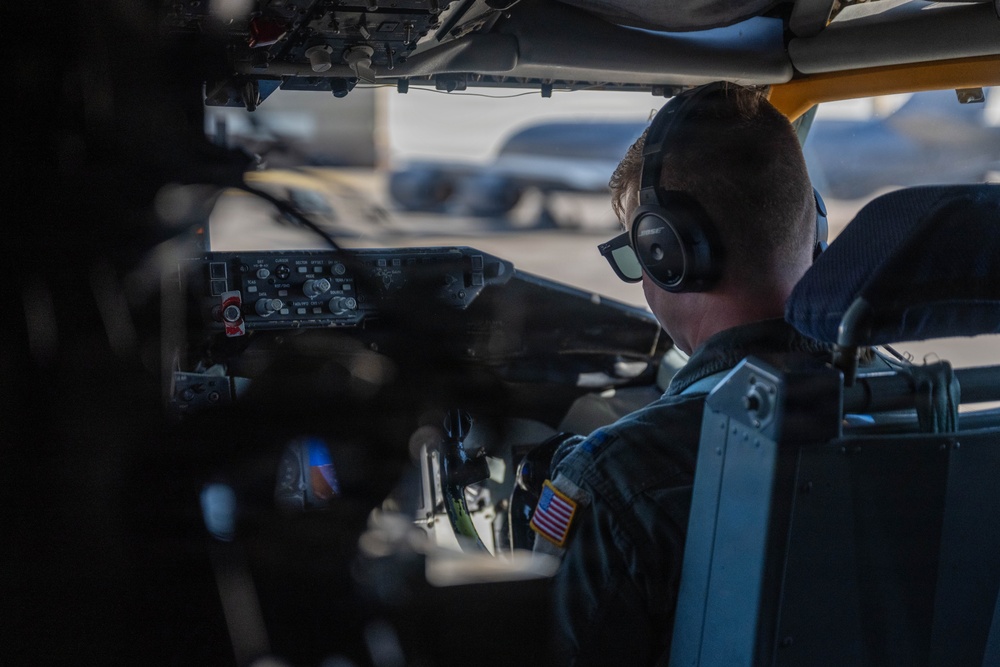 A military aviator is seated in the cockpit of an aircraft, viewed from behind and slightly to the side. The individual is wearing a flight suit with a U.S. flag patch, Bose aviation headphones, and sunglasses. The cockpit interior shows instrument panels and controls, with a flight line and other aircraft visible through the windscreen in the background.