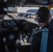 A military aviator is seated in the cockpit of an aircraft, viewed from behind and slightly to the side. The individual is wearing a flight suit with a U.S. flag patch, Bose aviation headphones, and sunglasses. The cockpit interior shows instrument panels and controls, with a flight line and other aircraft visible through the windscreen in the background.