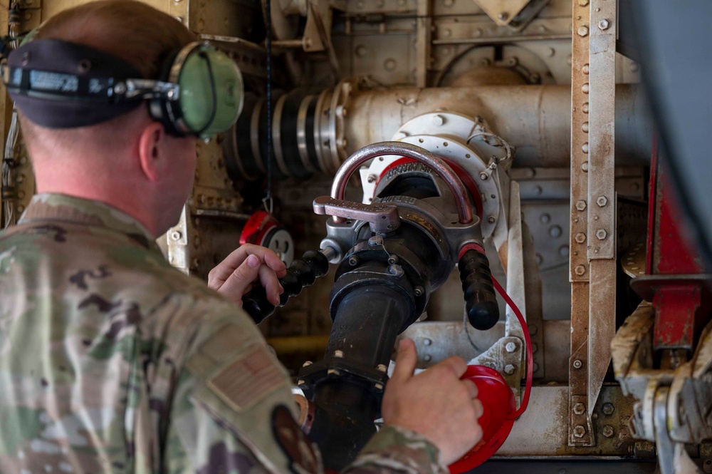 A military service member in MultiCam/OCP camouflage uniform is working on what appears to be aircraft fueling or hydraulic equipment. The individual is wearing hearing protection (green ear defenders) and a headlamp, and is manipulating a large fuel or fluid coupling/nozzle assembly. The setting appears to be inside or adjacent to a military aircraft, indicated by the riveted metal panels and mechanical components visible in the background.