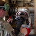 A military service member in MultiCam/OCP camouflage uniform is working on what appears to be aircraft fueling or hydraulic equipment. The individual is wearing hearing protection (green ear defenders) and a headlamp, and is manipulating a large fuel or fluid coupling/nozzle assembly. The setting appears to be inside or adjacent to a military aircraft, indicated by the riveted metal panels and mechanical components visible in the background.