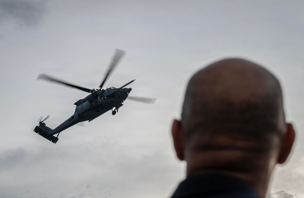 A military helicopter is captured in flight against an overcast sky, with a bald individual visible in the foreground out of focus. The helicopter appears to be a dark-colored military variant, possibly a Sikorsky UH-60 Black Hawk or similar model, with rotor blades in motion. The composition uses a shallow depth of field to contrast the observer in the foreground with the aircraft in the background.