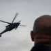 A military helicopter is captured in flight against an overcast sky, with a bald individual visible in the foreground out of focus. The helicopter appears to be a dark-colored military variant, possibly a Sikorsky UH-60 Black Hawk or similar model, with rotor blades in motion. The composition uses a shallow depth of field to contrast the observer in the foreground with the aircraft in the background.