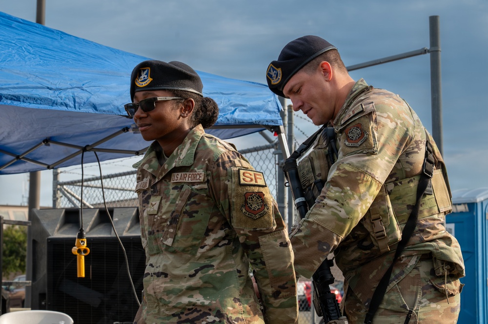 Two U.S. Air Force Security Forces (SF) personnel are pictured in OCP (Operational Camouflage Pattern) uniforms and black berets at what appears to be an outdoor post or checkpoint. The individual on the left is a female airman wearing sunglasses with a visible 'U.S. AIR FORCE' and 'SF' patch, while the male airman on the right appears to be checking equipment or a device. Both are equipped with tactical gear and appear to be on duty.