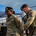 Two U.S. Air Force Security Forces (SF) personnel are pictured in OCP (Operational Camouflage Pattern) uniforms and black berets at what appears to be an outdoor post or checkpoint. The individual on the left is a female airman wearing sunglasses with a visible 'U.S. AIR FORCE' and 'SF' patch, while the male airman on the right appears to be checking equipment or a device. Both are equipped with tactical gear and appear to be on duty.