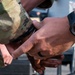 Close-up photograph of two individuals' hands, one wearing military camouflage (OCP/MultiCam pattern) uniform. One person is wearing a large digital Garmin Instinct Solar watch on their wrist. The hands appear to be in contact, possibly in a handshake, pulse check, or restraint scenario.