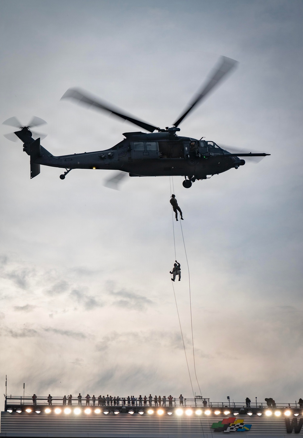 A military helicopter, appearing to be a Sikorsky UH-60 Black Hawk or similar variant, hovers over what appears to be a racing stadium or grandstand area. Two personnel are suspended on ropes beneath the aircraft, performing what appears to be a fast-rope or rappelling demonstration. Spectators are visible along the top of the grandstand structure below, watching the aerial display.