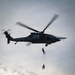 A military helicopter, appearing to be a Sikorsky UH-60 Black Hawk or similar variant, hovers over what appears to be a racing stadium or grandstand area. Two personnel are suspended on ropes beneath the aircraft, performing what appears to be a fast-rope or rappelling demonstration. Spectators are visible along the top of the grandstand structure below, watching the aerial display.