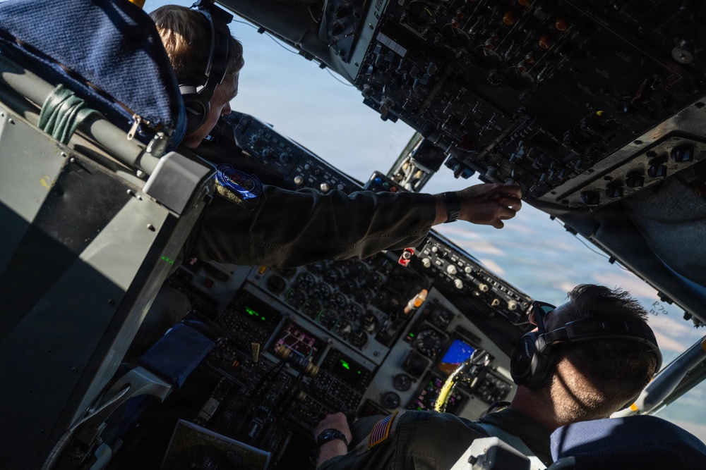 Two military aircrew members are visible in the cockpit of a large military aircraft, likely a tanker or transport aircraft such as a KC-135 or similar platform. One crew member is reaching up toward the overhead panel to manipulate controls or switches while the other observes. The aircraft appears to be in flight, with sky and clouds visible through the cockpit windows.