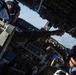 Two military aircrew members are visible in the cockpit of a large military aircraft, likely a tanker or transport aircraft such as a KC-135 or similar platform. One crew member is reaching up toward the overhead panel to manipulate controls or switches while the other observes. The aircraft appears to be in flight, with sky and clouds visible through the cockpit windows.
