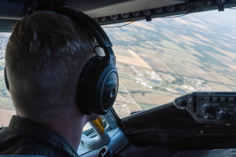 A pilot is seated in an aircraft cockpit, viewed from behind, wearing Bose aviation headphones and looking out the windshield at a patchwork of agricultural fields and a small town below. The aircraft appears to be at a moderate altitude during flight. Cockpit instrumentation and panels are partially visible on the right side of the frame.