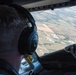 A pilot is seated in an aircraft cockpit, viewed from behind, wearing Bose aviation headphones and looking out the windshield at a patchwork of agricultural fields and a small town below. The aircraft appears to be at a moderate altitude during flight. Cockpit instrumentation and panels are partially visible on the right side of the frame.