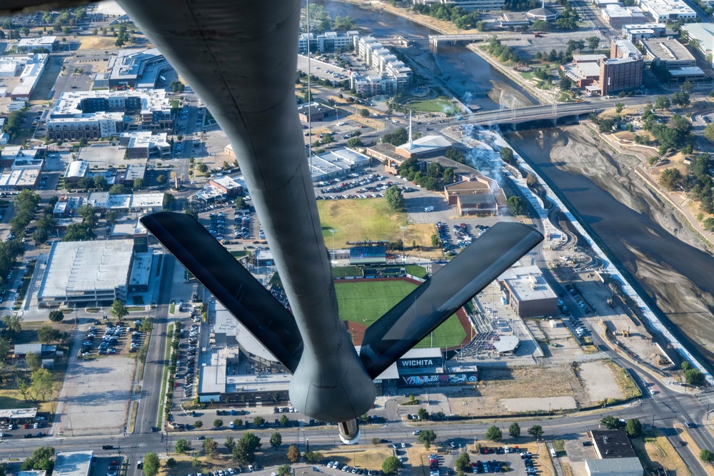 An aerial photograph taken from beneath a B-2 Spirit stealth bomber flying over downtown Wichita, Kansas. The aircraft's distinctive flying-wing shape and tail section are visible in the foreground, with the city's urban landscape, a baseball stadium, and the Arkansas River visible below. A 'WICHITA' sign is legible on the stadium, confirming the location.