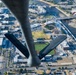 An aerial photograph taken from beneath a B-2 Spirit stealth bomber flying over downtown Wichita, Kansas. The aircraft's distinctive flying-wing shape and tail section are visible in the foreground, with the city's urban landscape, a baseball stadium, and the Arkansas River visible below. A 'WICHITA' sign is legible on the stadium, confirming the location.