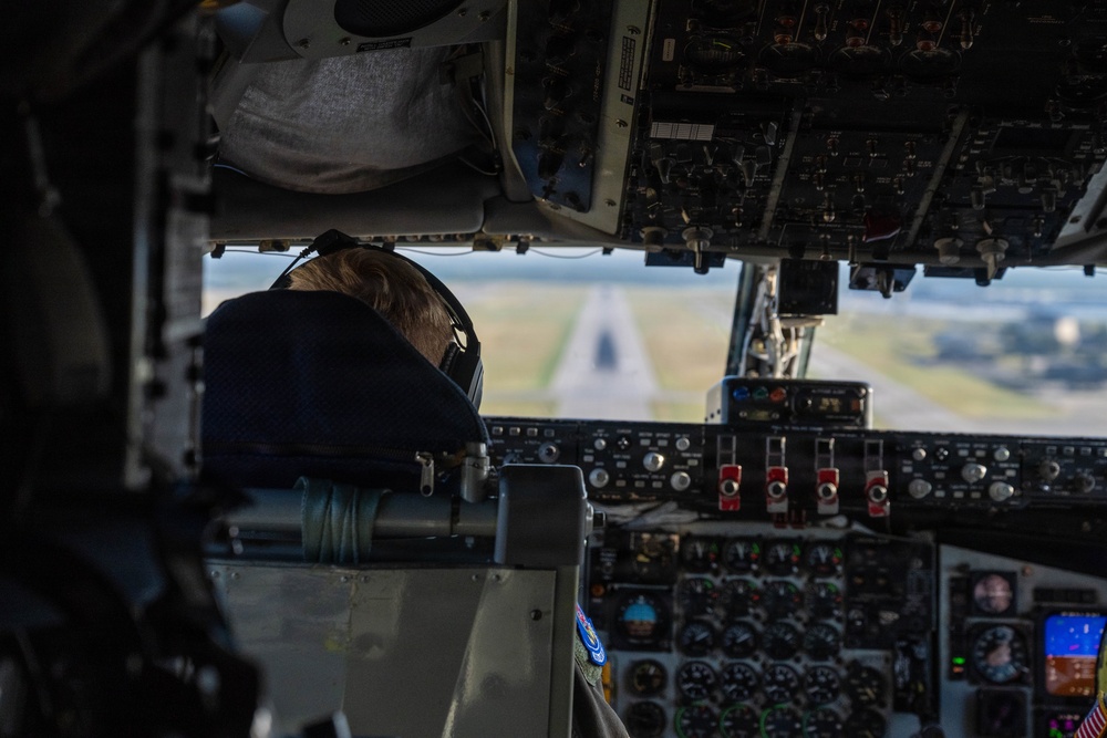 The image shows the interior of a military aircraft cockpit with a pilot visible from behind, wearing a flight cap with what appears to be a U.S. Air Force patch on their flight suit. The aircraft appears to be on final approach or low altitude flight, with a runway clearly visible through the windscreen. The cockpit is densely populated with analog and digital instruments, switches, and controls consistent with a large military transport or tanker aircraft.