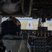 The image shows the interior of a military aircraft cockpit with a pilot visible from behind, wearing a flight cap with what appears to be a U.S. Air Force patch on their flight suit. The aircraft appears to be on final approach or low altitude flight, with a runway clearly visible through the windscreen. The cockpit is densely populated with analog and digital instruments, switches, and controls consistent with a large military transport or tanker aircraft.