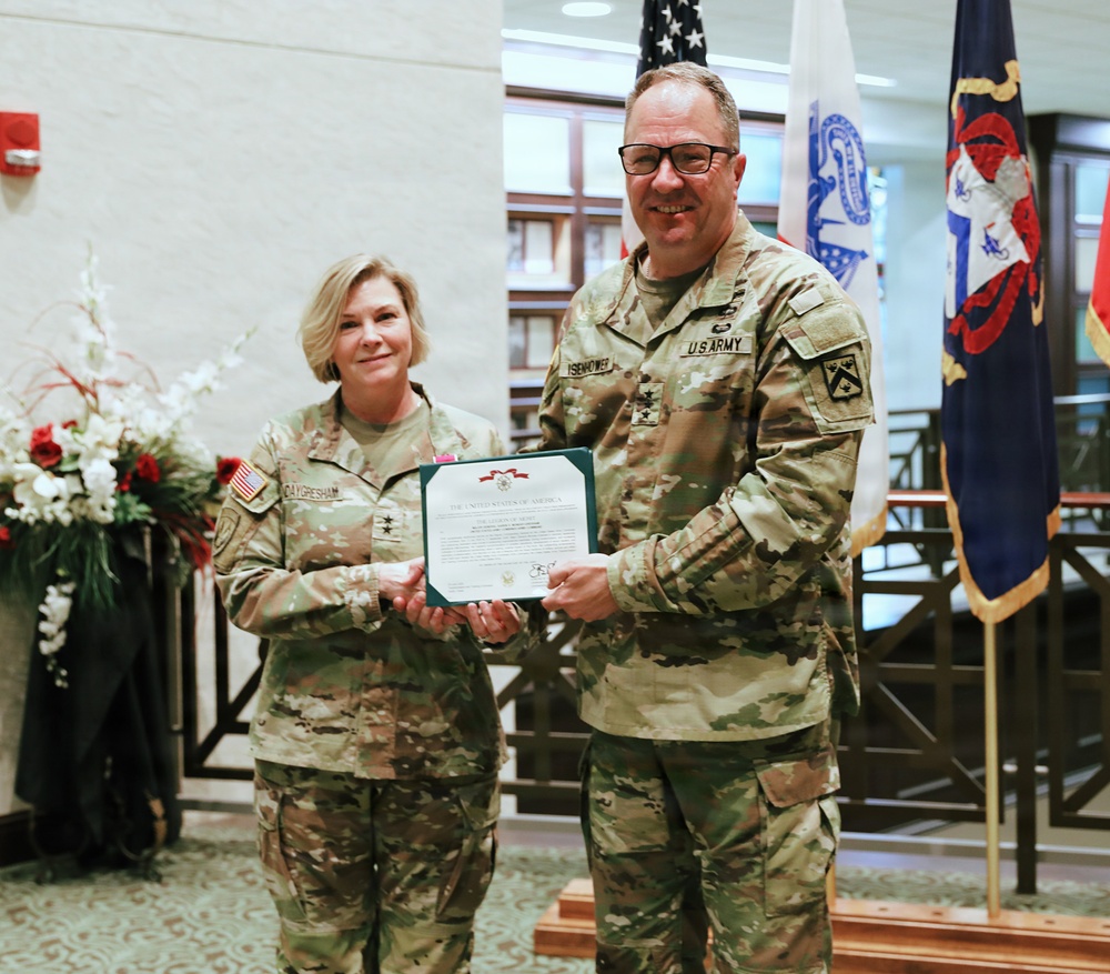 Two U.S. Army officers in OCP (Operational Camouflage Pattern) uniforms pose together holding a certificate during what appears to be a formal military award or recognition ceremony. The officer on the right, whose name tape reads 'ISENHOWER,' wears two stars indicating the rank of Major General, while the officer on the left, whose name tape reads 'DAYGRESHAM,' also appears to wear general officer rank insignia. The ceremony is taking place in an indoor military facility decorated with flags and floral arrangements.