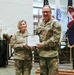 Two U.S. Army officers in OCP (Operational Camouflage Pattern) uniforms pose together holding a certificate during what appears to be a formal military award or recognition ceremony. The officer on the right, whose name tape reads 'ISENHOWER,' wears two stars indicating the rank of Major General, while the officer on the left, whose name tape reads 'DAYGRESHAM,' also appears to wear general officer rank insignia. The ceremony is taking place in an indoor military facility decorated with flags and floral arrangements.