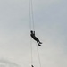 Two individuals in military-style uniforms are suspended from ropes against an overcast sky, performing what appears to be a rappelling or fast-rope descent exercise. Both figures are silhouetted against the grey sky, with one positioned higher than the other on separate rope lines. The activity is consistent with military or tactical training operations.
