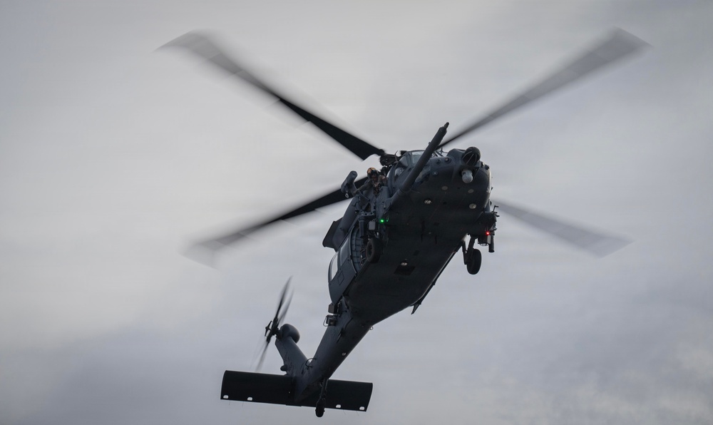 A dark gray military helicopter is captured in flight against an overcast sky. The helicopter is viewed from a slightly below-front angle, with its main rotor blades visibly blurred due to rotation. The aircraft appears to be a utility/special operations variant based on its configuration and dark tactical paint scheme.