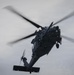A dark gray military helicopter is captured in flight against an overcast sky. The helicopter is viewed from a slightly below-front angle, with its main rotor blades visibly blurred due to rotation. The aircraft appears to be a utility/special operations variant based on its configuration and dark tactical paint scheme.