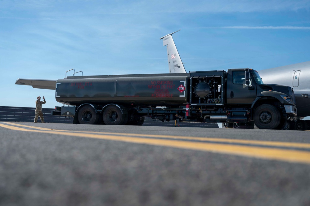 A military fuel tanker truck is positioned on an airfield tarmac alongside a large military aircraft, likely a KC-135 or similar tanker/transport aircraft. A uniformed service member is visible on the left side, appearing to direct or signal during a refueling operation. The image is taken from a low angle near the tarmac surface, emphasizing the scale of the fuel truck.