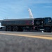 A military fuel tanker truck is positioned on an airfield tarmac alongside a large military aircraft, likely a KC-135 or similar tanker/transport aircraft. A uniformed service member is visible on the left side, appearing to direct or signal during a refueling operation. The image is taken from a low angle near the tarmac surface, emphasizing the scale of the fuel truck.